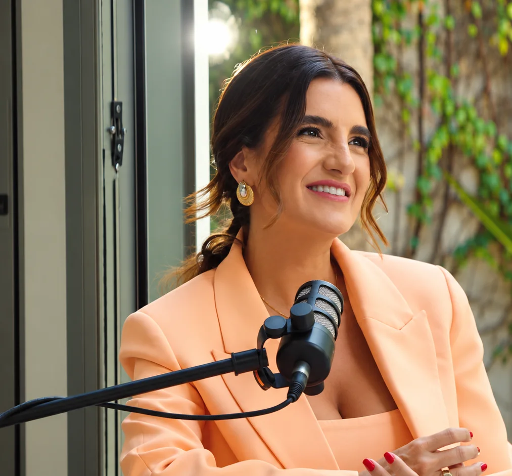 Ana Lisboa sorrindo ao microfone, de blazer salmão, junto a uma janela